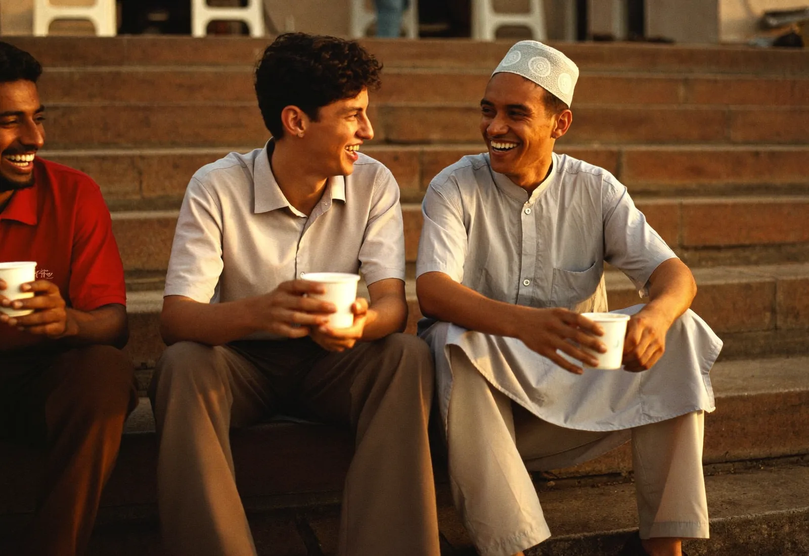 Three men sitting on stone steps at golden hour holding cups of coffee, laughing together
